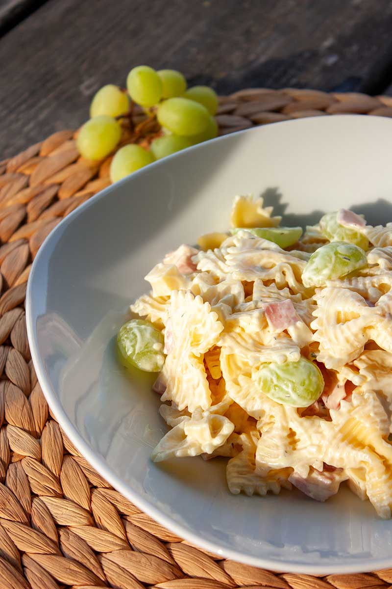 Wit bord met pastasalade met ham, kaas en witte druiven. Naast het bord ligt een tros witte druiven